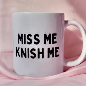May include: White ceramic mug with the words "MISS ME KNISH ME" printed in bold black letters. The mug has a curved handle and sits on a pink fabric background. The mug is a standard size for coffee or tea.