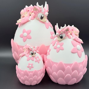 May include: Three decorative dragon egg sculptures in varying sizes. The eggs are white with pink flower accents and a pink dragon figure on top. The base of each egg is pink and petal-shaped.