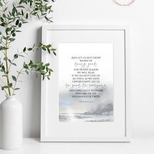 Könnte beinhalten: Gerahmter Druck mit dem Text "Und lasst uns nicht müde werden, Gutes zu tun..." in einem weißen Rahmen. Das Kunstwerk zeigt eine Aquarelllandschaft in Blau- und Grautönen. Links steht eine weiße Vase mit grünen Zweigen.