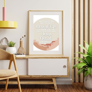 May include: Framed print with the text "And this is the way to have eternal life..." in a modern design. The print features a neutral color palette with beige, peach, and white tones. It is displayed on a wooden cabinet.
