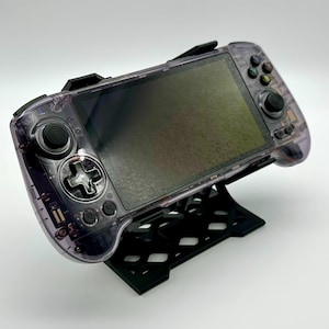May include: A transparent purple handheld gaming console with black buttons and a black stand. The console has a screen and control buttons on both sides. The stand is black and has a geometric design.