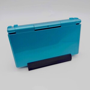 May include: Teal-colored handheld gaming console resting on a black stand. The console is closed, showing its smooth, glossy surface. The stand provides support, creating a clean, modern aesthetic. A classic gaming device.