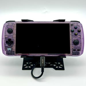 May include: A purple handheld gaming console with a black screen and a black stand. The console has a black and white logo on the front and a black cable connected to a black adapter.