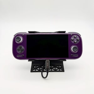 May include: A purple handheld gaming console with a black screen, analog sticks, and buttons, resting on a black geometric stand. A gray charging cable with the text AYN is plugged into the bottom center of the device, connecting to the stand.