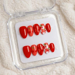 May include: A set of red press-on nails in a clear plastic case. The nails feature cherry and gingham designs. Some nails are solid red, while others have cherry accents or a white and red checkered pattern.