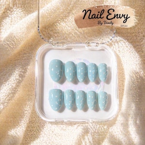 May include: A set of light blue press-on nails with white polka dots, arranged in a clear plastic case. The nails have an oval shape. The case is open, revealing the full set of nails. The text "Nail Envy By Trinity EST. 2023" is visible.