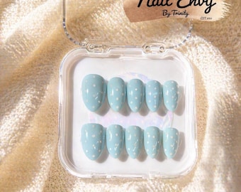 Pode incluir: Um conjunto de unhas postiças azuis claras com bolinhas brancas, dispostas em uma caixa de plástico transparente. As unhas têm formato oval. A caixa está aberta, revelando o conjunto completo. O texto "Nail Envy By Trinity EST. 2023" é visível.