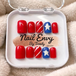 May include: A set of press-on nails in a clear case. The nails feature a patriotic design with red, white, and blue colors. Some nails are solid red, others have white stripes, and some have a white star on a blue background. The text "Nail Envy By Trinity EST. 2025" is visible.