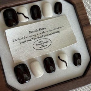 May include: A set of press-on nails in a wooden tray. The nails are a mix of glossy black and off-white, with one nail featuring a black curved design. A tag reads "French Pairs" and includes text about the brand.