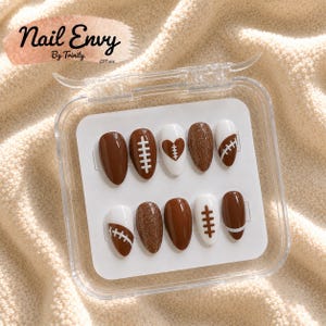 Puede incluir: Un juego de diez uñas postizas en forma de almendra en un estuche de plástico transparente. Los diseños presentan colores marrón y blanco con motivos de fútbol americano, incluyendo cordones blancos, un corazón marrón y detalles de purpurina. Texto Nail Envy By Trinity EST.2021.