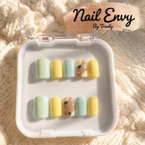 May include: A set of press-on nails in a clear plastic case. The nails feature various designs, including pastel green with yellow stripes, solid yellow, pastel green, and a brown nail with a lemon design. The text "Nail Envy By Trinity EST. 2023" is visible.