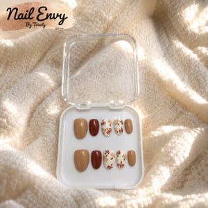 May include: A set of ten press-on nails in a clear plastic case, featuring solid tan, solid dark brown, and cream-coloured nails with brown and tan abstract leaf patterns. The text Nail Envy By Trinily EST 2023 is visible in the upper left corner.