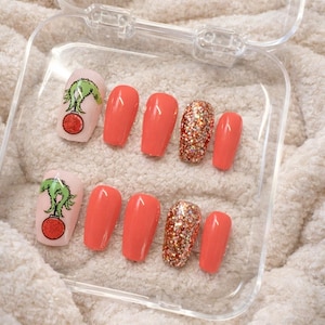 May include: A collection of press-on nails presented in a clear plastic case. The nails showcase a variety of designs: solid coral-coloured nails, glitter-covered nails, and nails with a green Grinch character design on a light pink background.