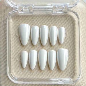 May include: A set of ten pearlescent white press-on nails in a clear plastic case. The nails are oval-shaped and vary in size. The case is open, revealing the nails arranged neatly on a beige surface. The nails are a beauty product.