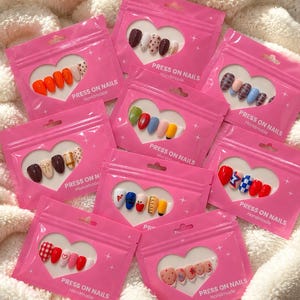 May include: A collection of eight pink plastic pouches, each containing a set of press-on nails with various designs, colors, and patterns. Each pouch features a heart-shaped clear window and the printed text PRESS ON NAILS Handmade.