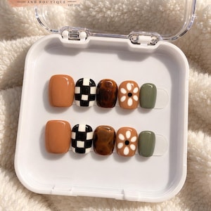 May include: A set of ten press-on nails in a white plastic case labeled Nail Envy By Trinity AND BOUTIQUE EST. 2025. The nails feature a glossy finish with designs including solid tan, black and white checkerboard, brown marble, white daisy flowers on tan, and solid olive green.