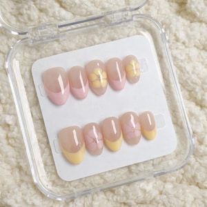 Puede incluir: Un juego de uñas artificiales en una caja de plástico transparente. Las uñas son de forma ovalada con base natural, puntas rosas y amarillas y diseños florales. Las uñas están dispuestas sobre una tarjeta blanca.