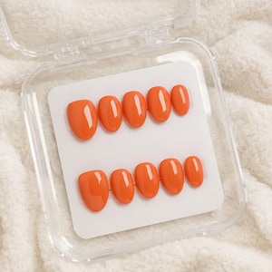 May include: A set of orange press-on nails in a clear plastic case. The nails are oval-shaped and arranged in two rows of six on a white card. The case is open, revealing the glossy, solid-colored nails.