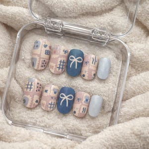 Puede incluir: Un juego de uñas postizas en una caja de plástico transparente. Las uñas presentan varios diseños, incluyendo patrones de sellos postales, lazos y purpurina. Los colores incluyen azul claro, azul marino y beige.