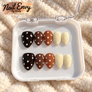 May include: A set of ten press-on nails in a white plastic case, featuring a gradient of brown and cream shades with white polka dots. The text Nail Envy By Trinity AND BOUTIQUE EST. 2023 is printed in black on the upper left corner of the case.
