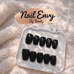 May include: A set of glossy black press-on nails in a clear plastic case. The case is open, revealing ten nails arranged in two rows. The top of the case features the text "Nail Envy By Trinity EST:2025" in black script.