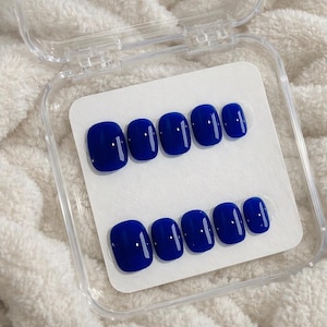 May include: A set of glossy, royal blue press-on nails. The nails are arranged in two rows of six, presented on a white card inside a clear plastic case. The nails have a rounded shape and are ready to apply.