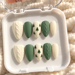 Op de afbeelding: Een set van tien amandelvormige plaknagels in een wit plastic doosje met het label Nail Envy By Trinity. De nagels hebben een mix van crème en saliegroene kleuren, texturen met kabelpatronen en kleine groene dennenboom-ontwerpen.