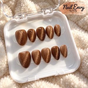 May include: A set of ten almond-shaped press-on nails in a glossy brown cat-eye finish with a shimmering gold magnetic effect. The nails are arranged in two rows inside a clear plastic storage case labeled Nail Envy By Trinity And Boutique.