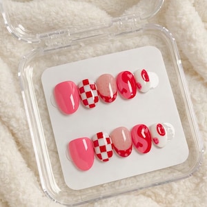 May include: A set of press-on nails in a clear plastic case. The nails feature a variety of designs, including solid pink, red and white checkered patterns, pink with red heart accents, and white with red lip designs.