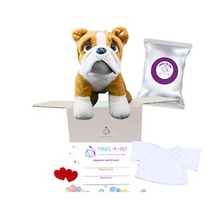May include: A plush bulldog toy with brown and white fur sits in an open cardboard box. The box contains a bag with a purple label, a white t-shirt, an adoption certificate, and two red heart decorations. The text "MAKE-A-PET" is visible.