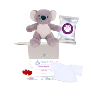 May include: A plush gray and white koala toy sits in an open cardboard box. Included are a silver foil packet with a purple logo, a white t-shirt, two small red felt hearts, and a paper adoption certificate with the text MAKE-A-PET printed on it.