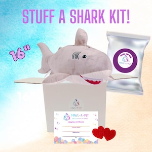 May include: A 16-inch plush shark toy in a box with a silver bag of stuffing, a Make-A-Pet adoption certificate, and two red felt hearts. The text reads STUFF A SHARK KIT!, 16, and Make-A-Pet. The shark is light gray with a white belly.