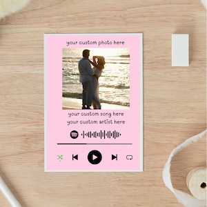 Puede incluir: Una tarjeta fotográfica rosa personalizada con una foto de una pareja en la playa. La tarjeta incluye campos de texto para una canción y un artista personalizados, junto con una interfaz de reproductor de música. La tarjeta está sobre una superficie de madera.