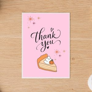 May include: A rectangular greeting card with a light pink background featuring the black cursive text thank you. Below the text is a slice of pumpkin pie topped with white whipped cream and a small orange pumpkin, surrounded by orange starbursts.