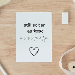 Può includere: Un biglietto di auguri bianco con il testo "still sober as f**k" e "I'm so so so proud of you" in scrittura nera. Un cuore nero è disegnato in basso. Un piccolo rettangolo bianco e una bobina di nastro sono visibili.