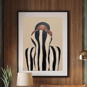 以下が含まれることがあります： 黒と白のストライプの衣服で顔を覆った人物が描かれた、額入りのアートプリント。アートワークは茶色の革張りのソファの上に掛けられています。ランプと植物が入った青い花瓶のあるサイドテーブルも表示されています。