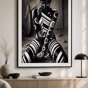 May include: A framed black and white wall art print featuring a person with dark skin covered in white painted geometric patterns, including circles and stripes, sitting in a cross-legged position. The artwork is displayed in a thin black frame.