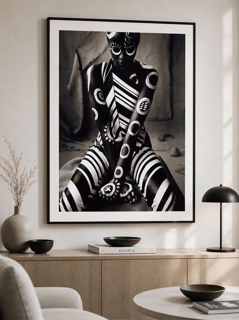 May include: A framed black and white wall art print featuring a person with dark skin covered in white painted geometric patterns, including circles and stripes, sitting in a cross-legged position. The artwork is displayed in a thin black frame.