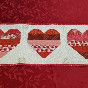 May include: A white table runner with three red, pink, and brown patchwork hearts. The runner has a red and white polka dot border.