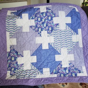 May include: A patchwork quilt with a purple border and a variety of blue and white patterns. The quilt features white crosses, blue geometric shapes, and patterned fabric with a chevron design. The quilt is approximately 40 inches square.