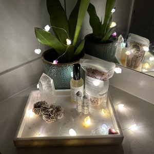 May include: A white marble tray with a clear quartz crystal, a rose quartz crystal, a brown selenite crystal, a bottle of essential oil, a bag of bath salts, and a string of fairy lights with white and coloured bulbs.