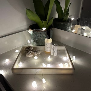 Peut inclure: Un plateau en marbre blanc avec une guirlande de lumières en cristal blanc, une petite bouteille d'huile essentielle et un cristal sur un comptoir blanc.
