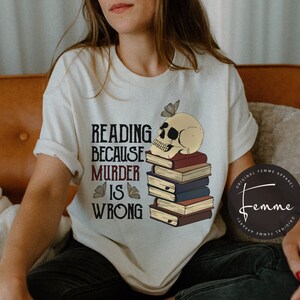 May include: White t-shirt with a skull wearing a stack of books and butterflies. The text on the shirt reads "Reading because murder is wrong."