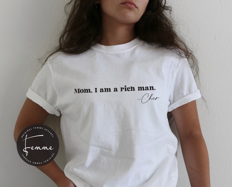 May include: White t-shirt with the text "Mom, I am a rich man. -Cher" printed on the front.