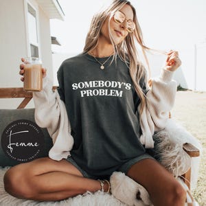 May include: A woman wearing a dark gray t-shirt with the text "Somebody's Problem" in white lettering. She is wearing a white cardigan and has her hair down. She is holding a glass of coffee.