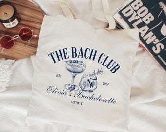 May include: A white canvas tote bag with blue text that reads "The Bach Club July 2024 Olivia's Bachelorette Austin, TX". The bag features two illustrations of cocktails.