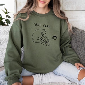 May include: A green sweatshirt with the text "SELF CARE" and a black and white illustration of a frog holding a cup of tea.