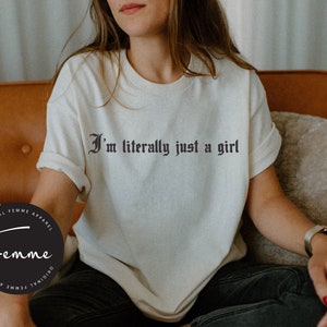 May include: A white t-shirt with the text "I'm literally just a girl" printed in black gothic font. The shirt is being worn by a person sitting on a couch. The text is centered on the shirt.