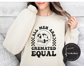 May include: A cream-colored sweatshirt with a black graphic print that reads "All Men Are Cremated Equal" with a skull and checkered pattern. The graphic is in a retro font style. The sweatshirt has a crew neck and long sleeves. The image also includes a logo for "Original Femme Apparel" in the bottom right corner.