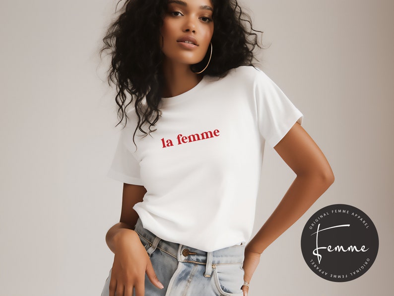 May include: White t-shirt with the text "la femme" in red lettering. The shirt is being worn by a person with long black hair. A black and white circle with the text "Original Femme Apparel" is in the bottom right corner of the image.